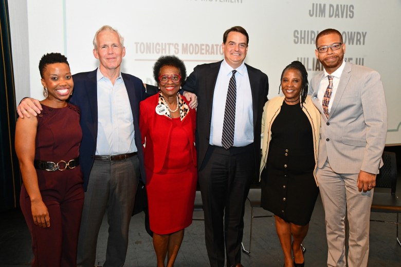 Six adults stand shoulder to shoulder on a stage, smiling toward the camera at a formal event. They are dressed in suits and dresses, with one person wearing a red outfit and a round pin. A projection screen behind them lists names and event information.