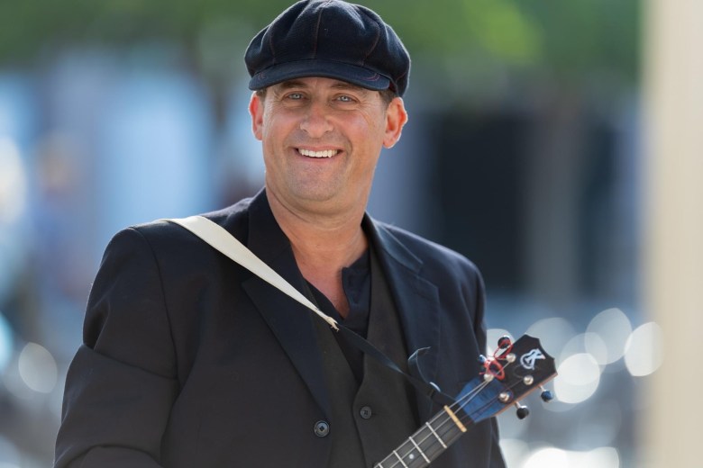 A medium close-up portrait of musician Lil' Rev, a middle-aged man with a warm, friendly smile and blue eyes. He is wearing a black flat cap (newsboy style), a black blazer over a matching vest, and a dark shirt. A white instrument strap across his shoulder holds a ukulele, of which only the headstock and part of the fretboard are visible. The background is softly blurred, suggesting an outdoor setting with dappled sunlight.