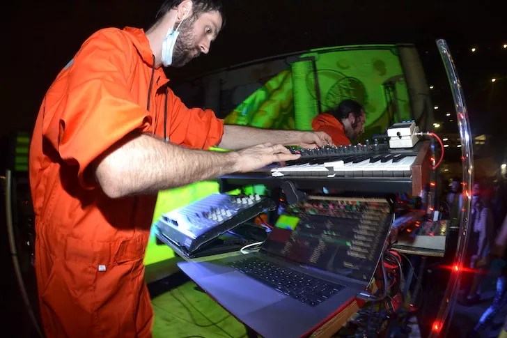 A musician in a bright orange jumpsuit and a white face mask tucked under their chin performs at a night event. They are focused on adjusting a synthesizer and mixer on a tiered stand, with a laptop displaying audio software in the foreground and green-tinted projections visible on the wall behind them.