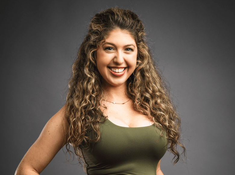 A studio portrait of a person with long, curly brown hair and a bright smile. They are wearing an olive green sleeveless top and a delicate gold necklace, posed against a solid dark grey background.