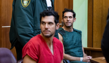 two handcuffed men, one in a red shirt and one in a navy kevlar vest, sit in a court room and look toward their right