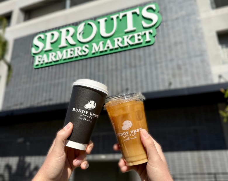 Two hands holding a hot coffee to-go cup and an iced coffee, both with buddy brew dog logos, cheers in front of a Sprouts Farmers Market