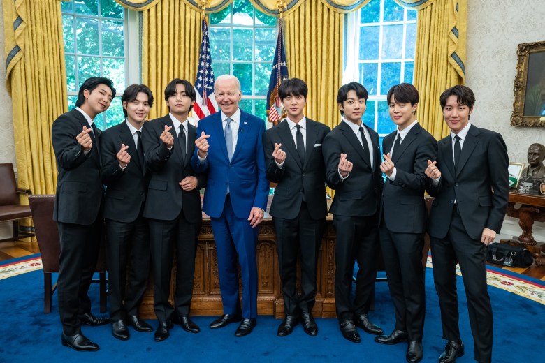 A professional group photograph taken in the Oval Office showing President Joe Biden standing with all seven members of the South Korean pop group BTS. Everyone is dressed in dark formal suits, and they are all performing the "finger heart" hand gesture.