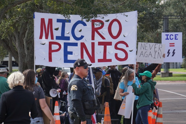 A crowd of people hold protest signs and a large banner reading “MICRO PENIS” during an outdoor demonstration, with a law enforcement officer standing nearby.