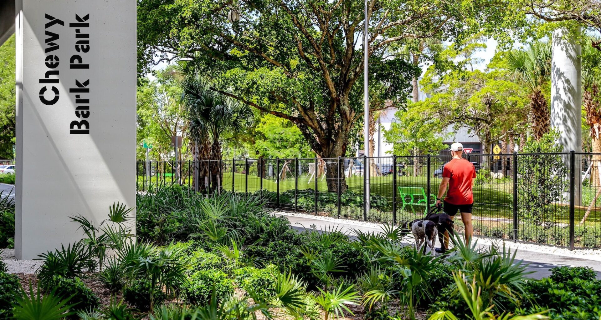 Photo of a person walking two dogs along a path under an overpass. A column below the overpass has a sign reading, "Chewy Bark Park"