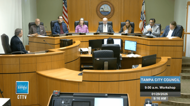 A wide shot of the Tampa City Council seated at a curved wooden dais during a morning workshop. Seven council members are visible behind the desk, with the City of Tampa seal and American and Florida flags centered on the wall behind them. A digital overlay in the corner identifies the meeting as a "Tampa City Council 9:00 a.m. Workshop" dated January 29, 2026.