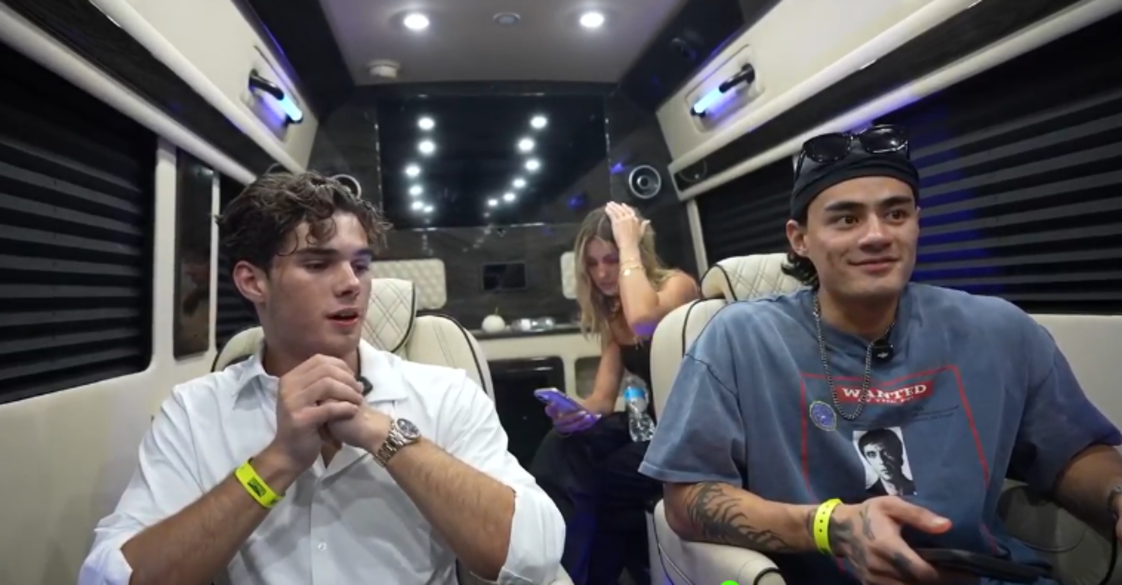 Clavicular sits on the left in a sprinter van. He is wearing a white-button down shirt. He is seated next to another man wearing a blue t-shirt. There is woman sitting behind them.