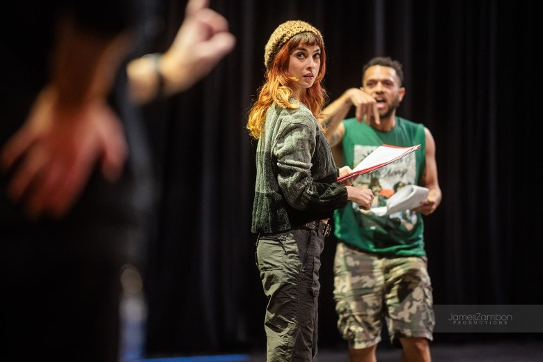A performer in a beanie and grey cargo pants stands on stage, looking back toward an out-of-focus figure in the extreme foreground. In the background, a second performer holding a script appears to be mid-dialogue against a backdrop of black curtains.
