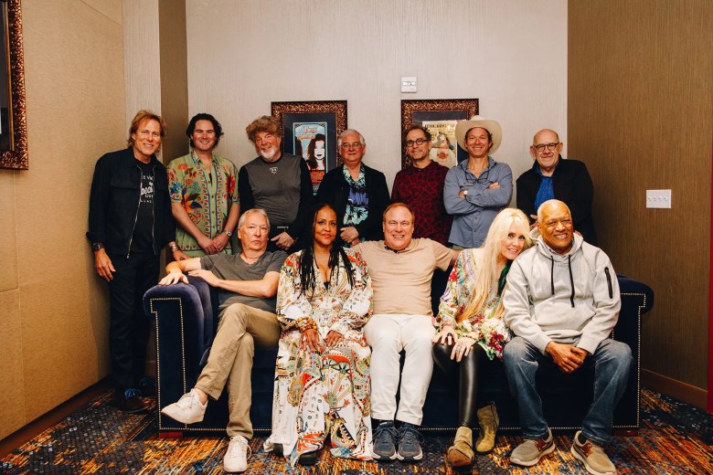welve members of the Coral Reefer Band pose together in a room with tan walls. Five people sit on a navy blue velvet sofa, while seven others stand behind them. The group is dressed in a variety of styles, including colorful patterned shirts, casual hoodies, and a wide-brimmed hat. Two framed posters hang on the wall in the background."