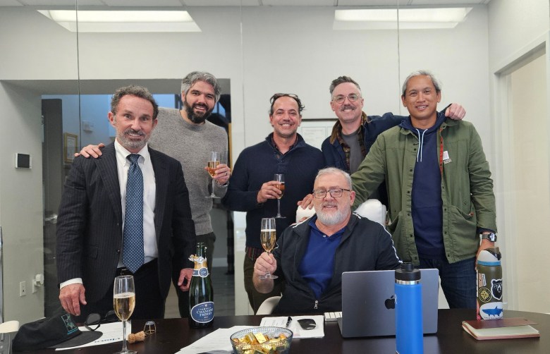 A group of seven people pose together in an office, smiling and holding glasses of sparkling wine around a desk with a laptop and documents.