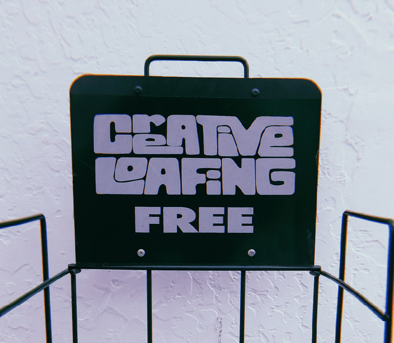 A black metal sidewalk sign reads “Creative Loafing Free,” mounted on a stand against a white stucco wall.