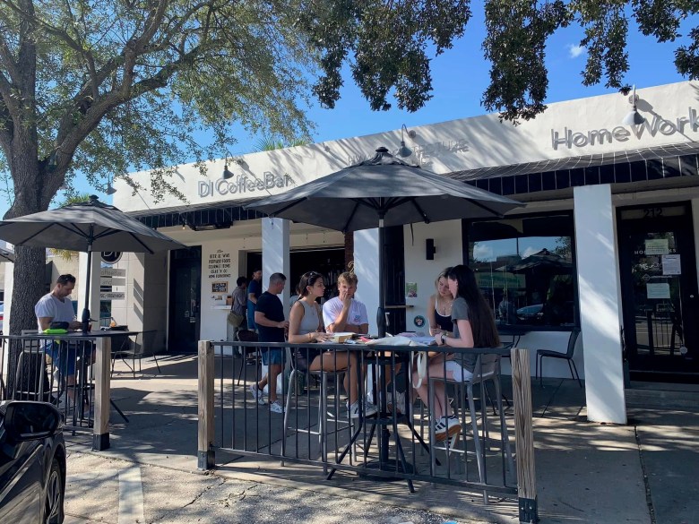 People sit at outdoor patio tables shaded by black umbrellas in front of "DI Coffee Bar" and an adjacent business labeled "HomeWork." The sunlit scene shows individuals engaged in conversation and looking at papers, while leafy trees cast shadows across the sidewalk and the white storefronts.
