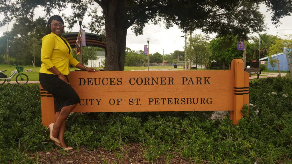 a woman in a yellow shirt stands in front of a wooden sign that reads "Deuces Corner Park"