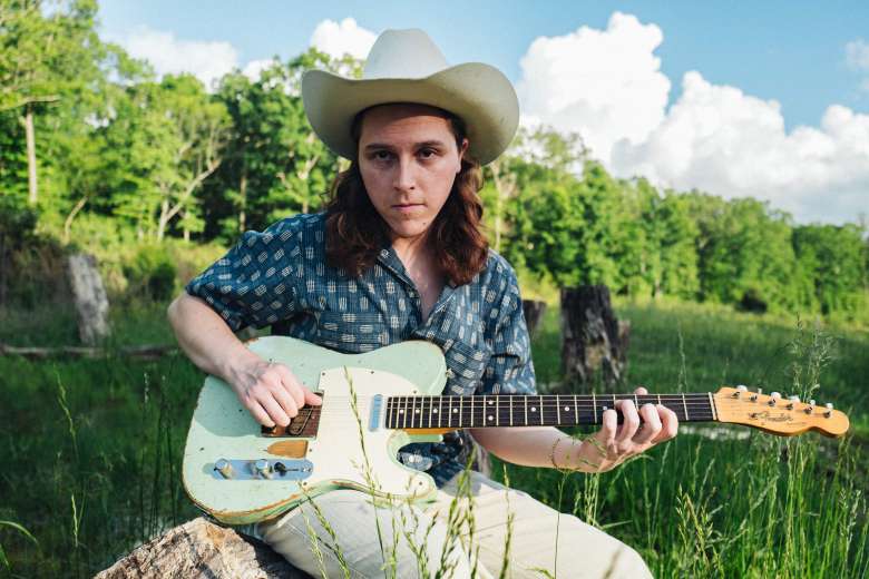 Musician Daniel Donato sits on a fallen log in a grassy field, wearing a white cowboy hat and a patterned blue shirt while playing a seafoam-green Fender Telecaster guitar.