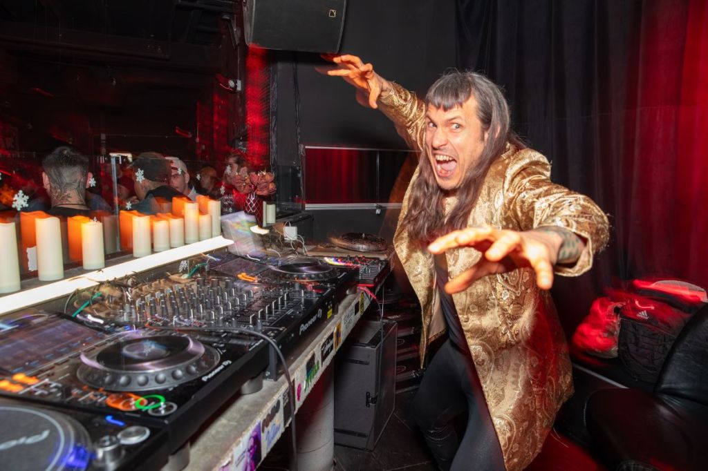Photo of a man next to CDjs wearing a golden coat. 