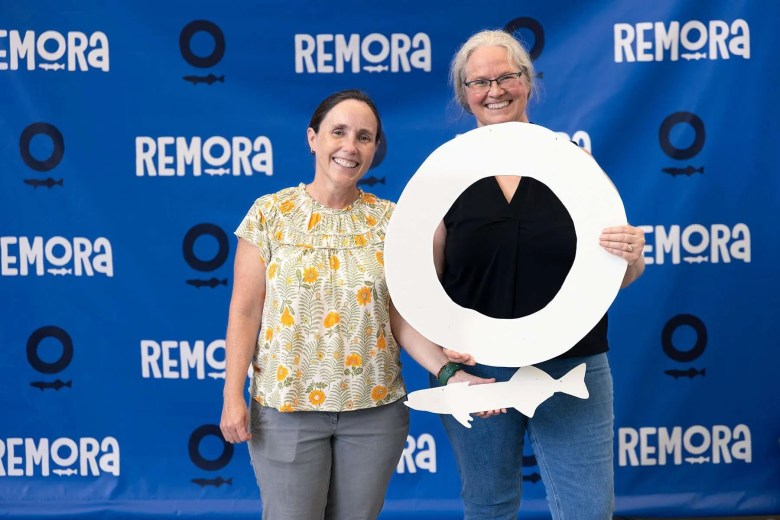 Two people stand side-by-side smiling in front of a blue backdrop patterned with the word "REMORA" and its logo. The individual on the left wears a yellow and white floral print blouse and grey trousers, while the individual on the right wears a dark sleeveless top and blue jeans. Together, they hold a large white cutout of the letter "O" and a white cutout silhouette of a remora fish.