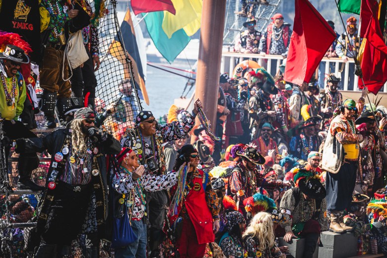 A large, energetic crowd of people dressed as pirates cheering and waving beads on the deck of a ship