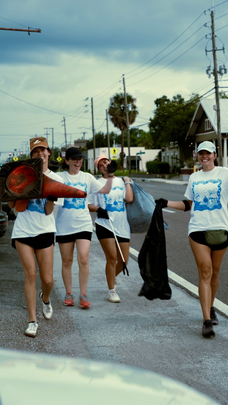Four people walk down a sidewalk smiling and laughing while carrying bags of litter and a discarded orange traffic cone. They are all wearing matching white t-shirts with a blue graphic, shorts, and baseball caps.