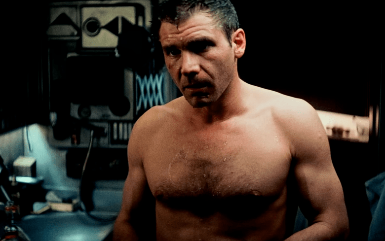 A high-contrast, moody film still featuring a man with a grit-covered face and damp skin. The lighting is low and atmospheric, casting deep shadows across his chest and the mechanical fixtures behind him, creating a sense of tension and exhaustion.