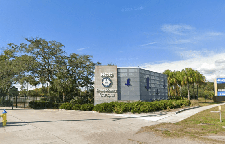 A sunlit outdoor view of the entrance to the Hillsborough Community College (HCC) Dale Mabry Campus. The main signage consists of a concrete wall featuring the HCC seal and a large, grey mesh structure adorned with blue hawk silhouettes. Green shrubs line the base of the sign, with a clear blue sky and several palm trees in the background.