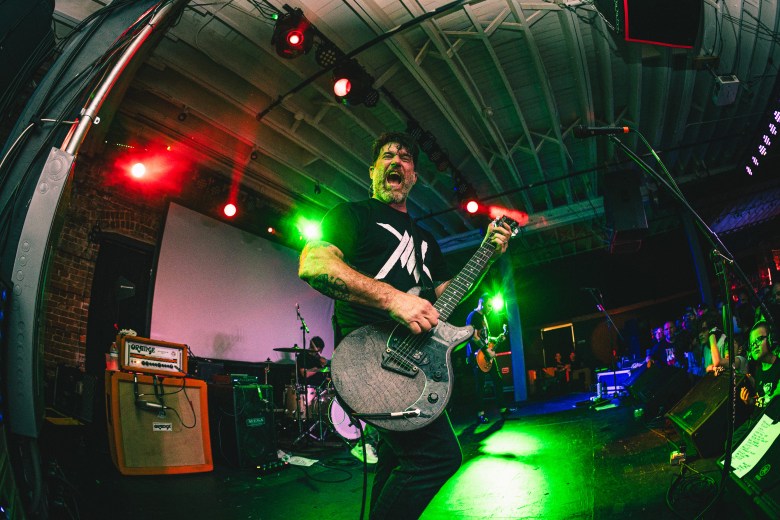Musician Chuck Ragan of Hot Water Music performs with intensity at Underbelly in Jacksonville. He is mid-shout while playing a black electric guitar, wearing a black t-shirt with a white graphic. The stage is captured with a wide-angle fisheye lens, illuminated by dramatic green and red stage lights.