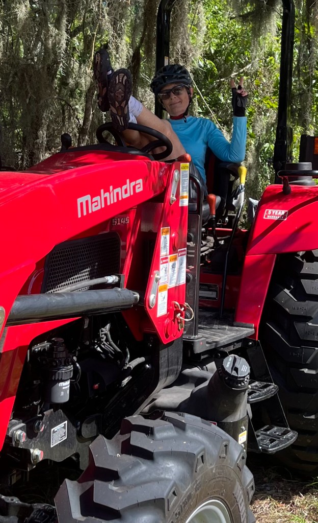 "A woman wearing a bicycle helmet and sunglasses leans back in the driver's seat of a red Mahindra tractor with her feet propped up on the steering wheel