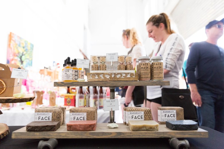 A close-up of a wooden display stand at an outdoor market featuring "Beard & Beauty" skincare products. The top shelf holds glass dropper bottles of face oil and small jars of face masks, while the bottom shelf displays several bars of artisanal soap in brown paper packaging labeled "FACE." In the bright, out-of-focus background, customers are seen browsing the various vendor stalls under a white canopy.