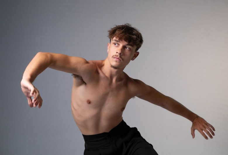 A shirtless individual with short, wavy brown hair and a mustache performs a contemporary dance pose against a neutral grey-to-white gradient background. They are wearing black high-waisted trousers, with their torso turned and arms extended in a fluid, expressive movement while looking off to the side.