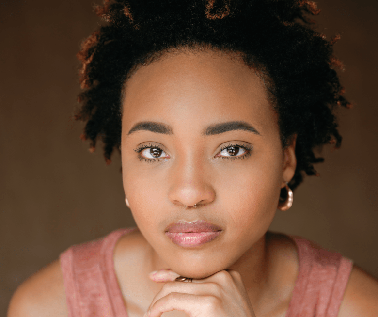 A serene studio portrait of a person with short, voluminous dark hair and golden-brown eyes looking directly at the camera. They are wearing a mauve sleeveless top and a small gold nose ring, with their chin resting on their hand, all set against a warm, out-of-focus brown background.