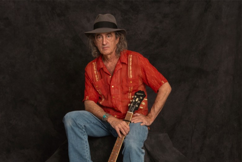 A studio portrait of Americana musician James McMurtry sitting against a dark, textured backdrop. He is wearing a grey fedora with a black band, a bright red short-sleeved button-down shirt with gold embroidered vertical panels, and blue jeans. He has long, greyish hair and wears a large turquoise cuff bracelet on his right wrist. He is holding the neck of an Epiphone acoustic guitar resting between his legs, looking directly at the camera with a neutral, steady expression.