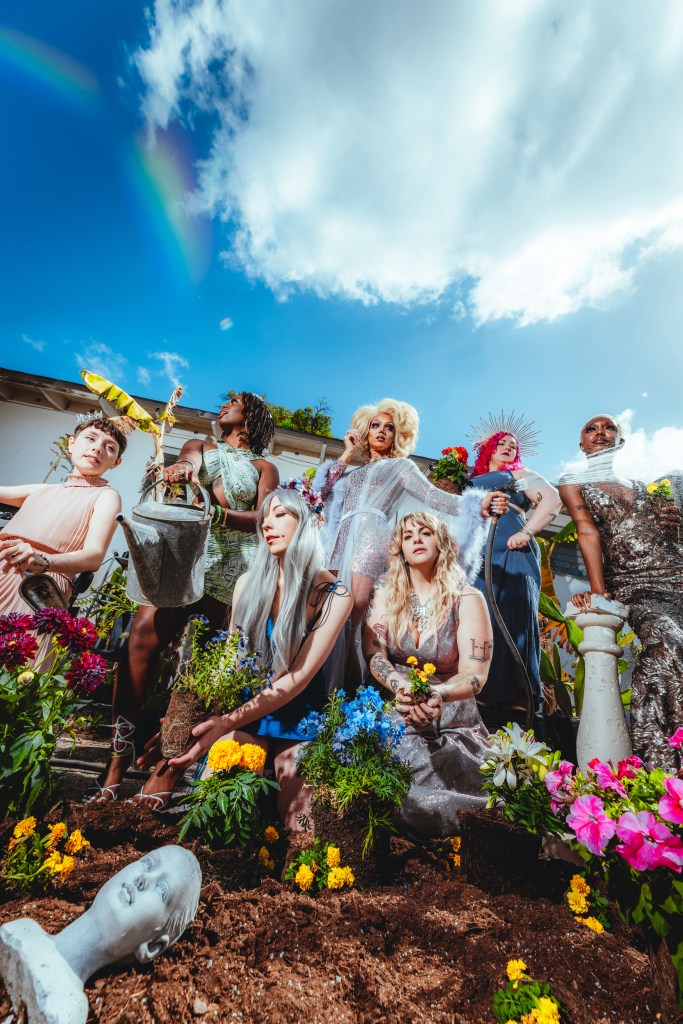 A high-angle, surreal group portrait of seven individuals in elaborate, shimmering costumes posed in a garden under a bright blue sky with a faint rainbow. The figures are engaged in various gardening activities; some hold watering cans or hoses, while others kneel in soil with flowers. The foreground features freshly turned earth and a white classical bust partially buried, with vibrant pink, yellow, and blue flowers scattered throughout the scene.