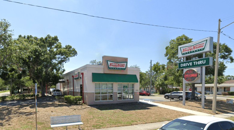 A Krispy Kreme doughnut shop with a green awning and drive-thru lane sits on a corner lot, with a roadside sign reading “Krispy Kreme Doughnuts” and “Drive Thru,” trees lining the street, and cars parked nearby on a sunny day.