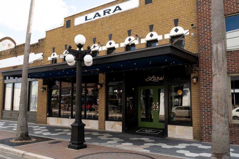 The exterior of "Lara Apothecary Bar & Bazaar," a two-story tan brick building with a black awning and green double doors. The storefront features large windows reflecting the street, a vintage-style black lamppost with five white globe lights in the foreground, and a geometric tiled sidewalk out front.