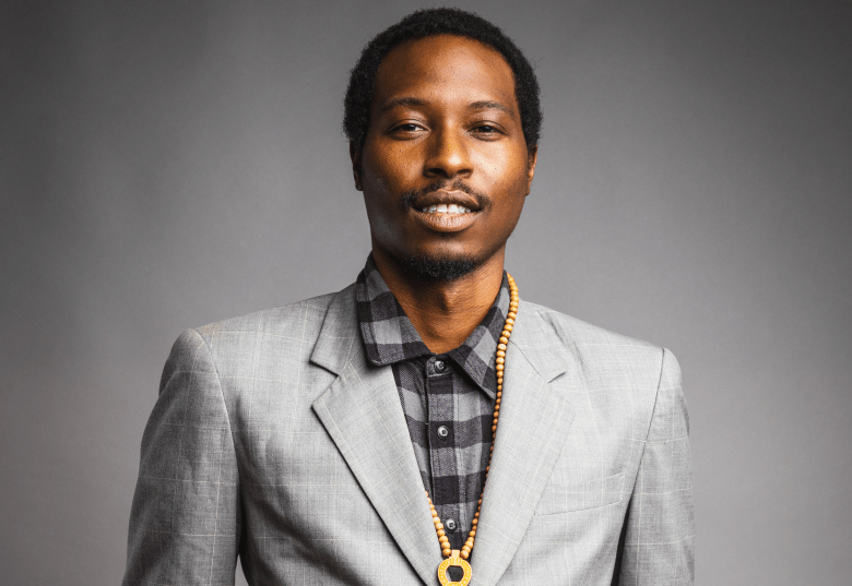 A studio portrait of a person with short dark hair and a mustache, looking directly at the camera with a slight smile. They are wearing a grey blazer over a black and grey plaid button-down shirt, accented with a long wooden bead necklace featuring a circular gold pendant, all set against a solid grey background.