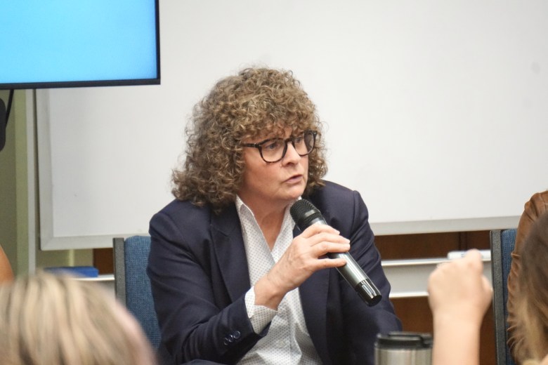 A person with curly brown hair and glasses sits in a blue chair, speaking into a black handheld microphone. They are wearing a dark navy blazer over a white patterned button-down shirt, captured in a candid moment during a panel or presentation with a white wall and a blue television screen in the background.