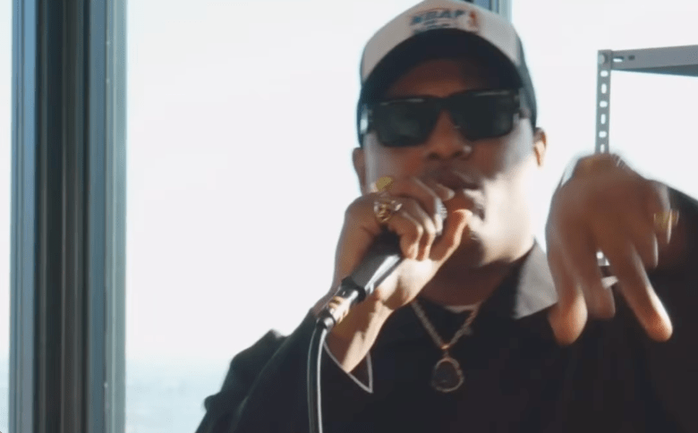 A close-up of a person wearing a black jacket, dark sunglasses, and a white and black baseball cap. They are holding a microphone to their mouth with a gold ring visible on their hand, gesturing with their other hand while standing in front of a bright window.