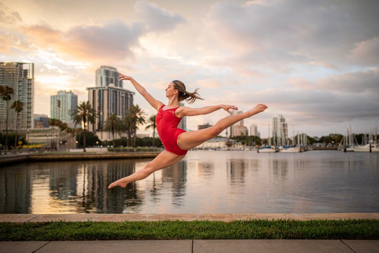 An individual in a red leotard performs a graceful leap in mid-air along a waterfront promenade at dusk. Their reflection is visible in the calm water, with a city skyline, palm trees, and sailboats in a marina under a soft, cloudy sky in the background.