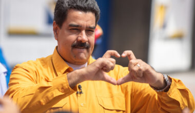 A mustachioed man wearing a yellow button down shirt makes a heart with his hands.
