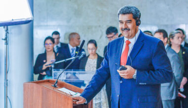 A man with grey hair stands before a podium in a blue suit.