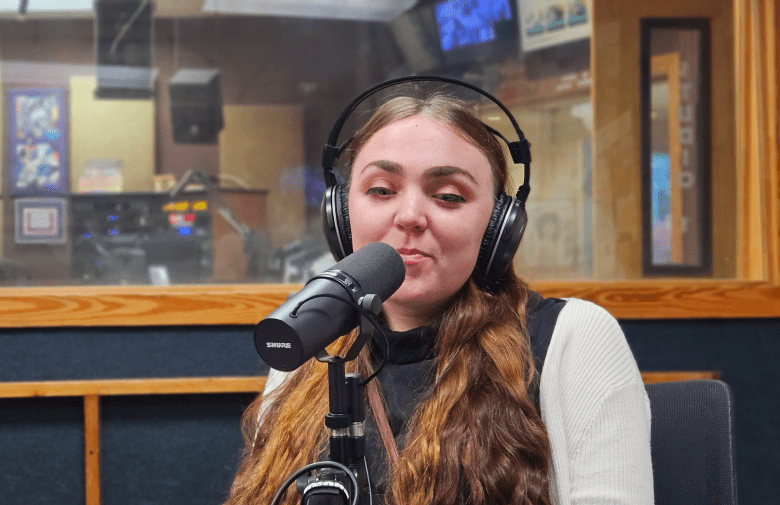 A close-up of a person with long, wavy brown hair wearing large over-ear headphones and speaking into a professional Shure microphone. They are dressed in a black top and a white cardigan, seated in a radio or podcast studio with a soundproof window and technical equipment visible in the blurred background.