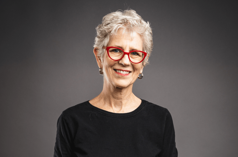 A studio portrait of a person with short, curly grey hair and a bright smile. They are wearing vibrant red cat-eye glasses, silver hoop earrings, and a black crew-neck top, posed against a solid grey background.