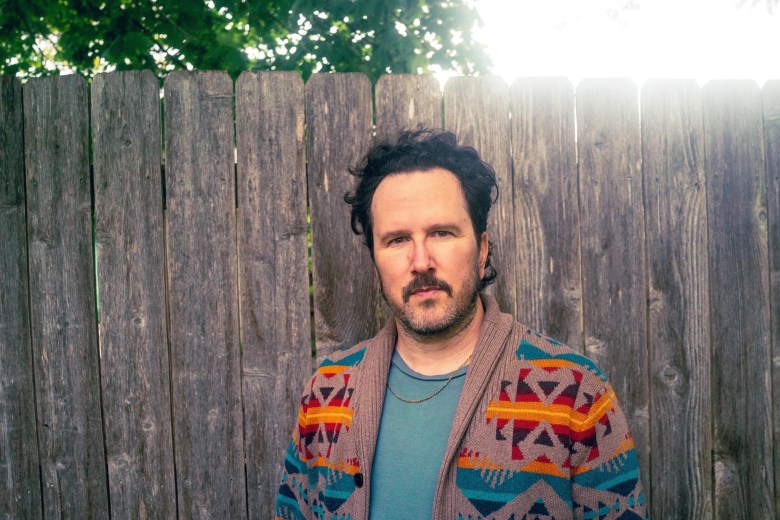 Musician Mason Jennings stands in front of a weathered wooden fence. They have dark, curly hair and a mustache, wearing a teal t-shirt under a tan cardigan with a vibrant geometric pattern in orange, red, and blue. Sunlight filters through trees in the background.