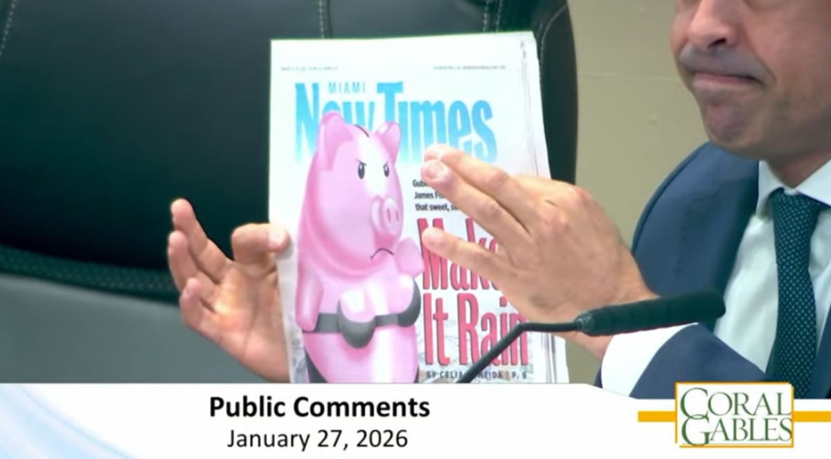 Commission meeting screenshot of Mayor Lago holding the cover of Miami New Times with four fingers. You can only see the bottom half of his face including his mouth and nose. The cover shows an angry pig wearing a bra and underwear.