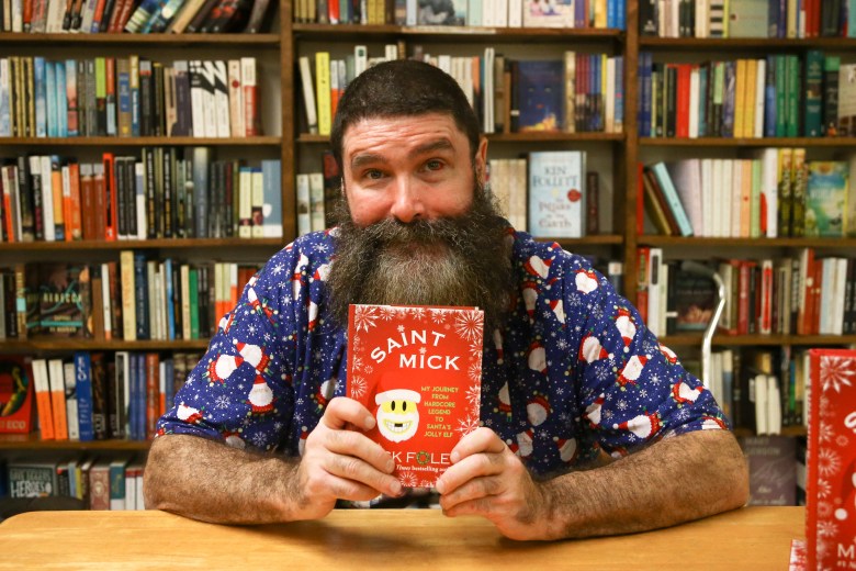 A close-up of author and former wrestler Mick Foley holding a copy of his book, "Saint Mick: My Journey From Hardcore Legend to Santa's Jolly Elf," during a signing event. He is wearing a blue shirt with a festive Santa pattern.