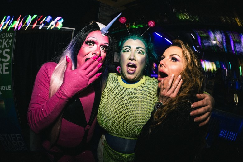 A close-up group shot captured with a wide-angle lens, creating a distorted, high-energy effect. The subjects are dressed in vibrant extraterrestrial costumes and makeup, standing in a dimly lit nightclub. A glowing neon "PHOTOS" sign and streaks of colorful light from a digital screen create a festive, party atmosphere in the background.