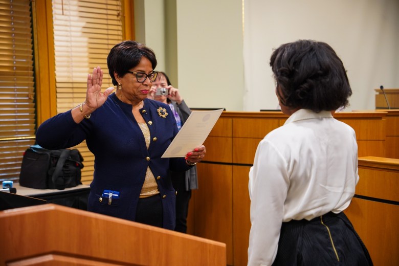 An indoor shot of a formal induction or oath-taking ceremony. The primary subject, wearing glasses and gold jewelry, reads from a certificate while performing a traditional hand gesture of affirmation. In the background, an observer is partially visible holding a small digital camera to capture the moment.