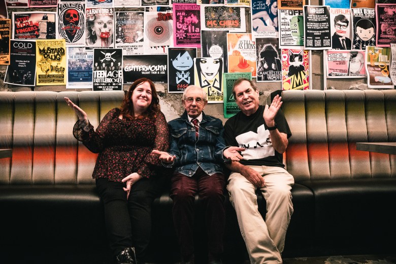 Three people are seated together on a long bench with a colorful, sunset-patterned backrest. Behind them, the wall is covered with a dense collage of colorful concert and event posters.