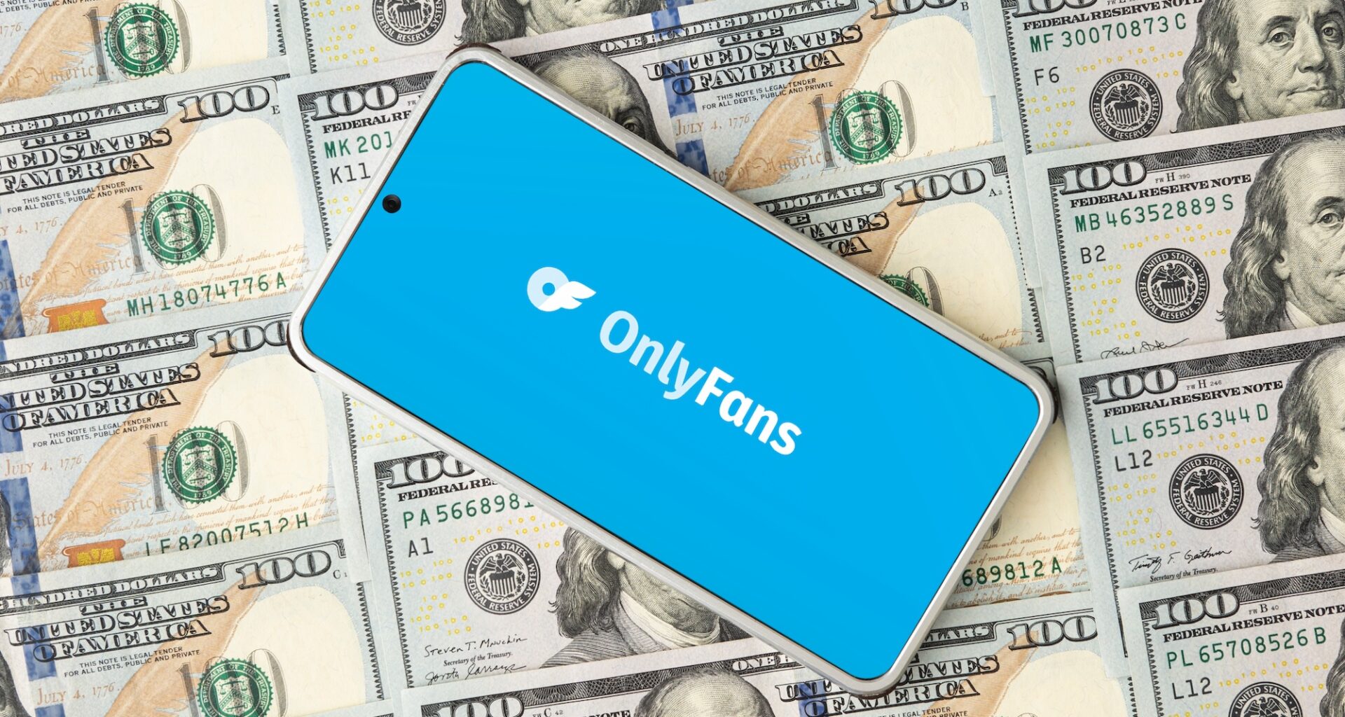 Photo of a smartphone on top of stacks of hundred-dollar bills. The phone displays the OnlyFans logo