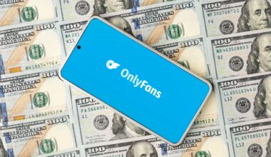 Photo of a smartphone on top of stacks of hundred-dollar bills. The phone displays the OnlyFans logo