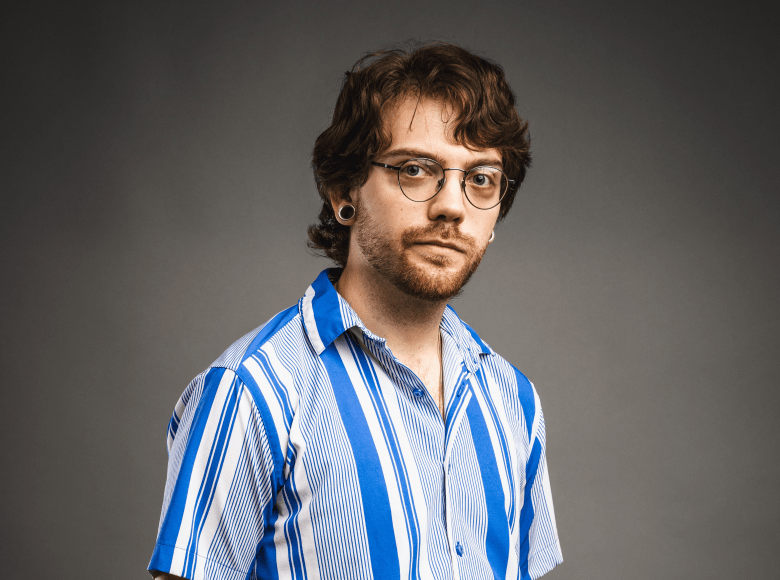 A studio portrait of a person with wavy brown hair, a beard, and glasses, looking directly at the camera with a neutral expression. They are wearing a blue and white vertically striped button-down shirt and have black gauge earrings, all set against a solid grey background.